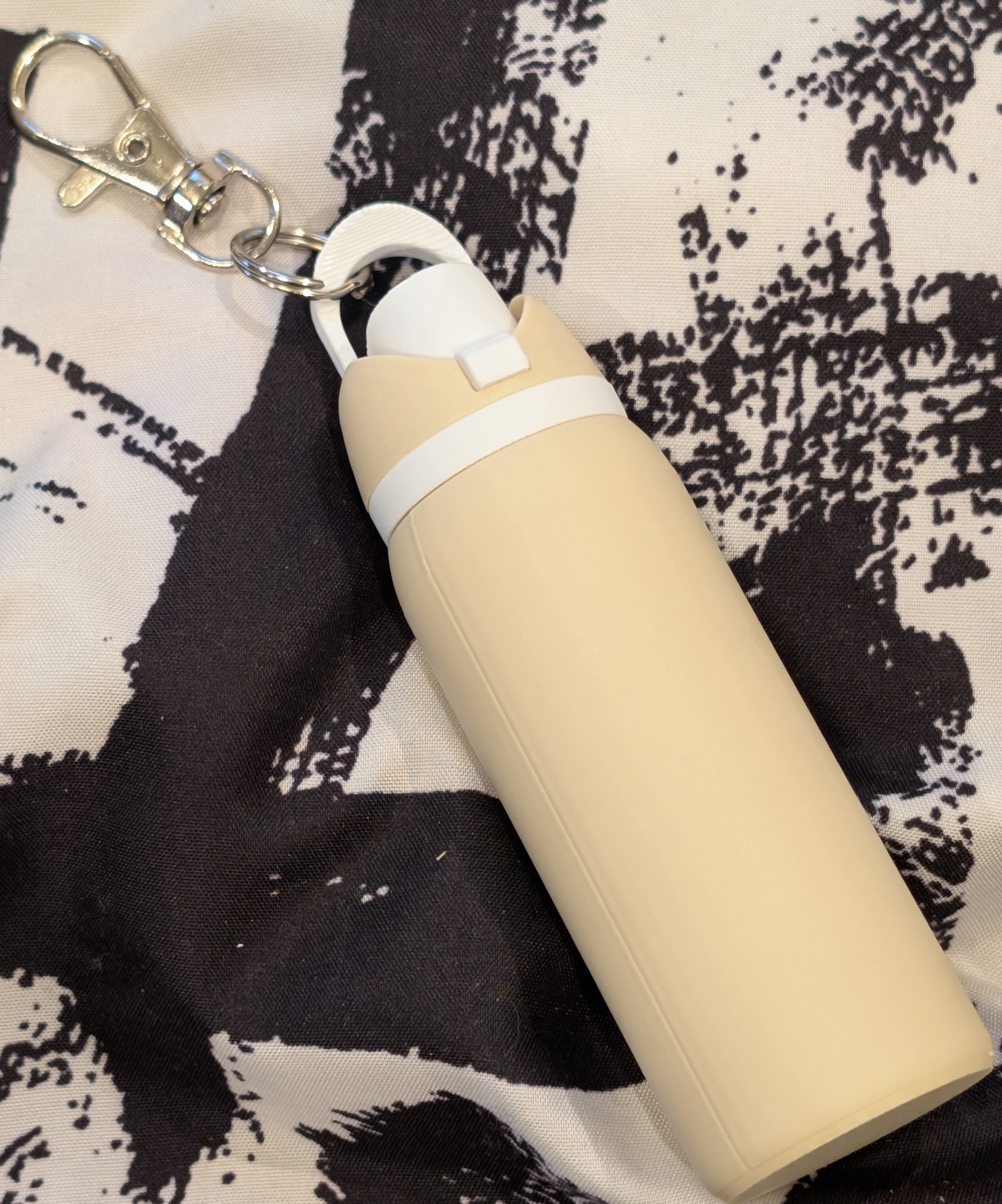 Beige water bottle with white cap and keychain on a patterned fabric background